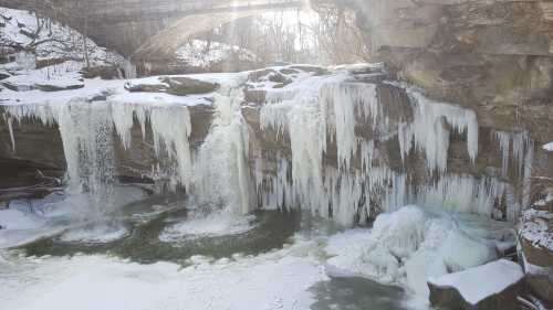 A frozen waterfall with icicles, surrounded by snow and rocky cliffs, under a bridge in a winter landscape.