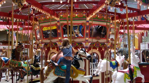 A child rides a carousel with colorful horses, surrounded by lights and festive decorations.