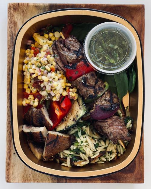 A meal box featuring grilled steak skewers, corn salad, orzo, and a side of green sauce.