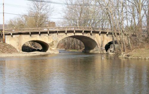 A stone bridge with two arches spans a calm river, surrounded by bare trees and a clear sky.