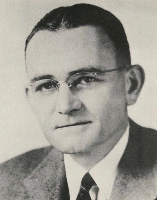 Black and white portrait of a man in a suit and tie, wearing glasses, with a neutral expression.