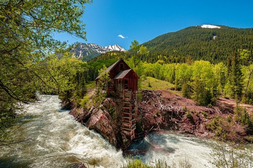 A rustic wooden cabin perched on a rocky outcrop by a rushing river, surrounded by lush greenery and mountains.