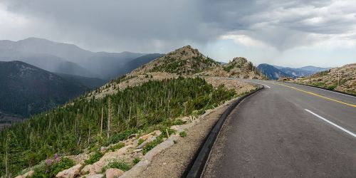 A winding road curves along a mountainous landscape under a cloudy sky, with lush greenery and distant peaks visible.