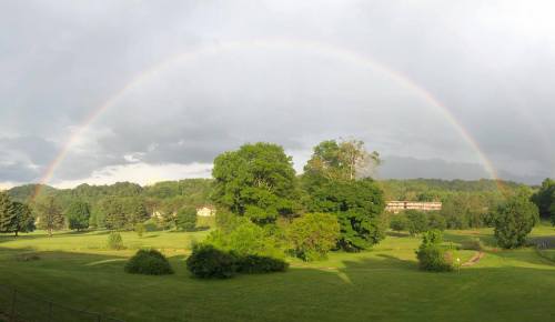 A vibrant rainbow arcs over a lush green landscape with trees and a distant building under a cloudy sky.