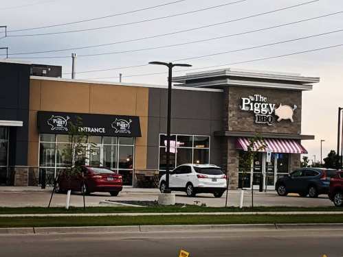 Exterior view of The Piggy BBQ restaurant, featuring a sign and outdoor seating area, with cars parked nearby.