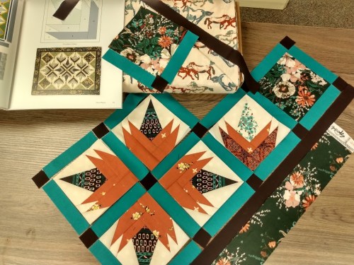 A colorful quilt layout featuring floral and geometric patterns, with a quilting book and fabric in the background.