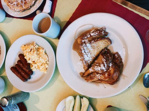 A breakfast spread featuring French toast, scrambled eggs, sausage links, and a cup of coffee on a colorful table.