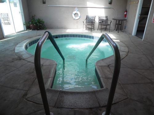 A small indoor hot tub with steps, surrounded by a concrete floor and chairs, illuminated by natural light.