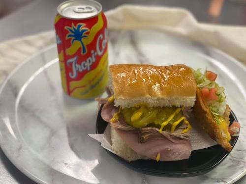 A sandwich with ham, mustard, pickles, and lettuce on a plate, next to a can of Tropi-Cool drink.