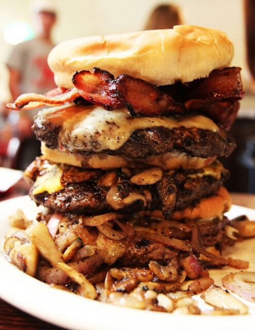 A towering burger stacked with bacon, cheese, and sautéed mushrooms, served on a plate.