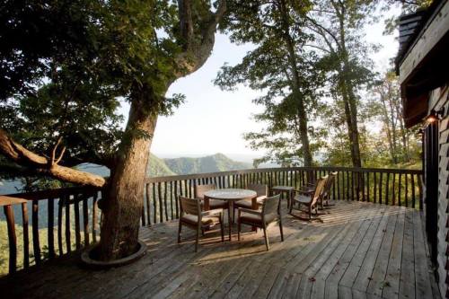 A wooden deck with a table and chairs, surrounded by trees and overlooking a scenic mountain view.