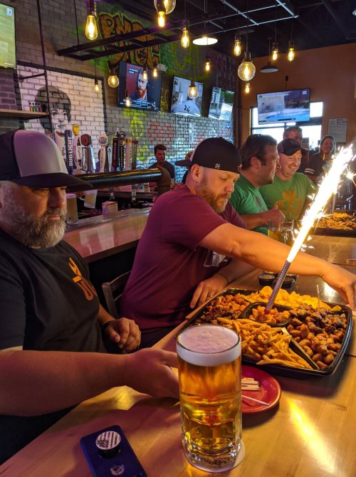 A group of friends at a bar enjoying a large platter of food with a sparkler, and a beer in the foreground.