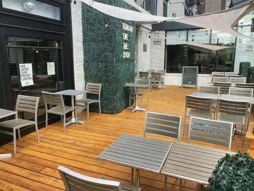 Outdoor dining area with wooden flooring, metal tables, and chairs, surrounded by greenery and a shaded canopy.