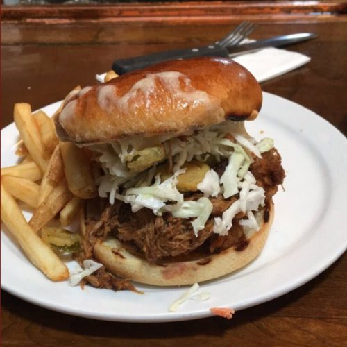 A pulled pork sandwich topped with coleslaw and pickles, served with a side of fries on a white plate.