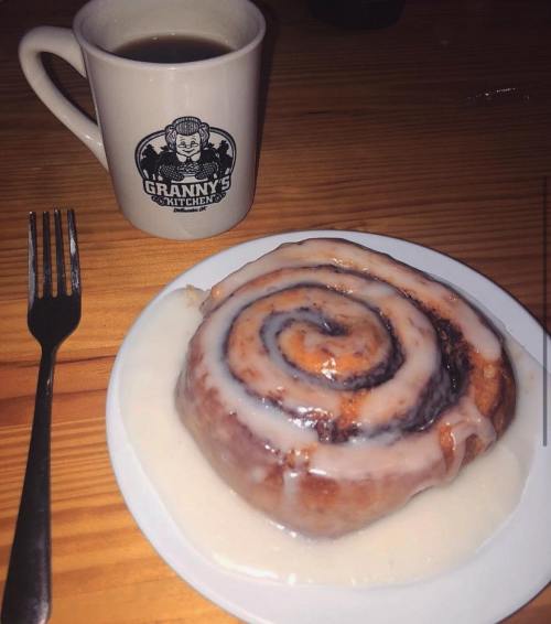 A cinnamon roll with icing on a plate next to a coffee mug labeled "Granny's Kitchen." Fork beside the plate.