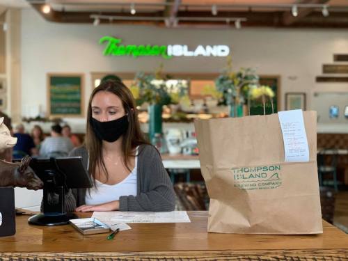 A masked employee at a restaurant takes an order, with a takeout bag and menu visible on the counter.