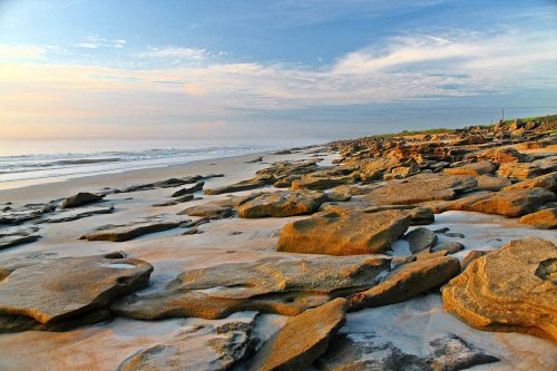 Rocky shoreline at sunrise, with gentle waves lapping against the sand and a clear blue sky above.