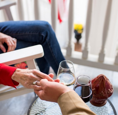 A close-up of two hands holding each other, with glasses of wine on a table and a cozy porch setting in the background.