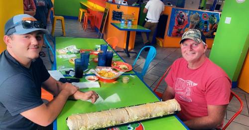 Two men sit at a colorful table in a restaurant, smiling beside a large burrito and plates of food.