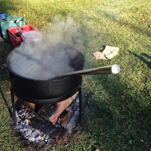 A large pot on a fire pit, with steam rising, set on grass. Nearby items include boxes and containers.