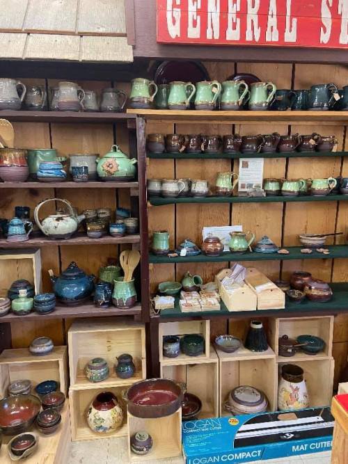 A display of various colorful pottery and ceramic pieces on wooden shelves in a general store.