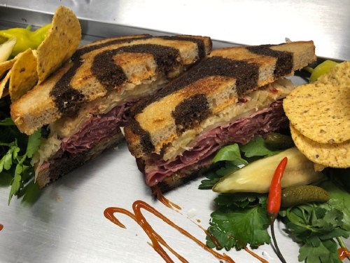 A close-up of a sliced sandwich with dark rye bread, filled with meat and sauerkraut, garnished with chips and pickles.