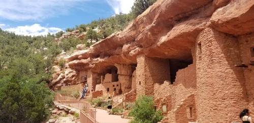 Ancient cliff dwellings nestled in a rocky landscape, surrounded by greenery and blue skies. Visitors explore the site.