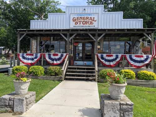 A rustic general store with a wooden facade, decorated with red, white, and blue bunting, surrounded by flowers.