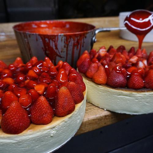 Two cheesecake layers topped with fresh strawberries and glossy red glaze, with a bowl of sauce in the background.