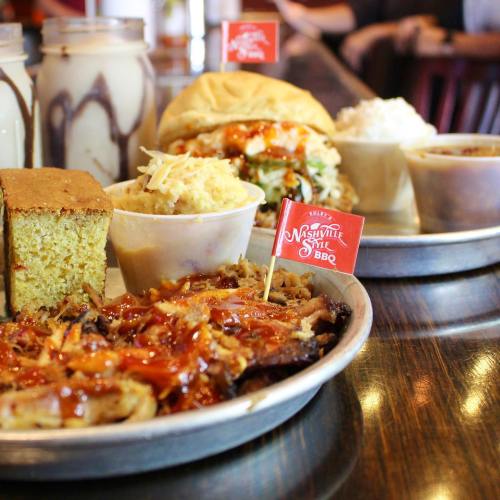 A platter of Nashville-style BBQ featuring pulled pork, cornbread, coleslaw, and a sandwich, with sauces and sides.