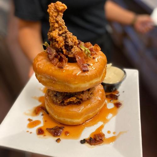 A stack of two glazed donuts topped with fried chicken, bacon bits, and drizzled with syrup, served on a white plate.