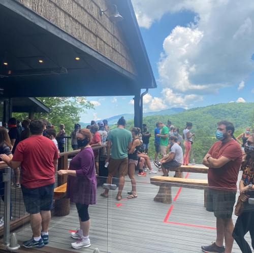 A crowd of people wearing masks waits in line outside a building with a scenic view of mountains and blue skies.