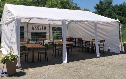 A white tent with wooden tables and chairs, labeled "The Angler," set up outdoors among greenery.