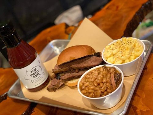 A BBQ meal featuring a brisket sandwich, baked beans, macaroni and cheese, and a bottle of barbecue sauce on a tray.