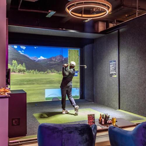 A golfer swings a club in a virtual golf simulator, with a scenic backdrop displayed on a large screen.