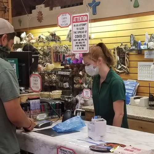 A cashier wearing a mask assists a customer at a store with various items displayed in the background.
