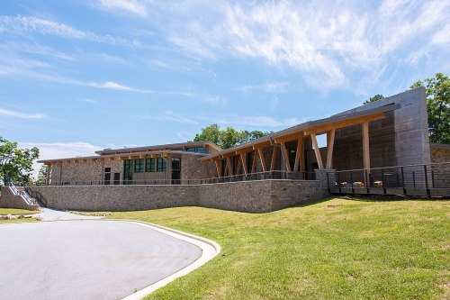 Modern building with stone and wood design, surrounded by greenery under a clear blue sky.