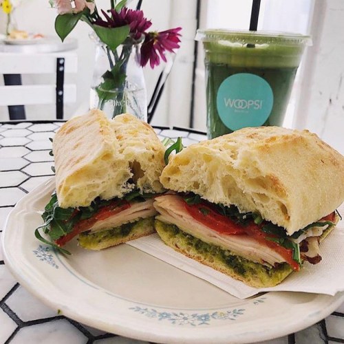 A fresh sandwich with turkey, arugula, and tomatoes on a plate, alongside a green smoothie and a small vase of flowers.