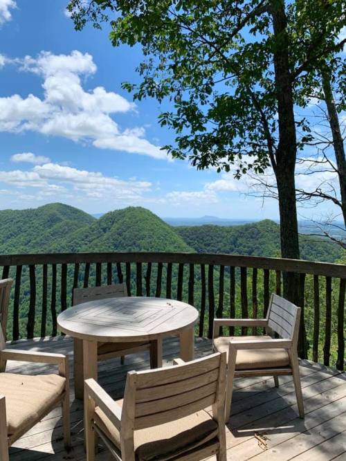 A wooden deck with a round table and chairs overlooks lush green mountains under a blue sky with fluffy clouds.