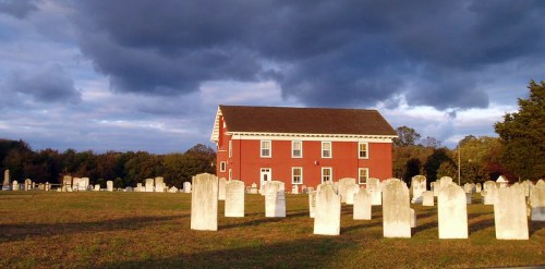A red building stands beside a cemetery with white gravestones under a cloudy sky.