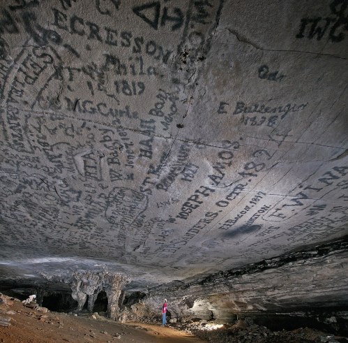A person stands in a cave, surrounded by walls covered in historical inscriptions and graffiti.