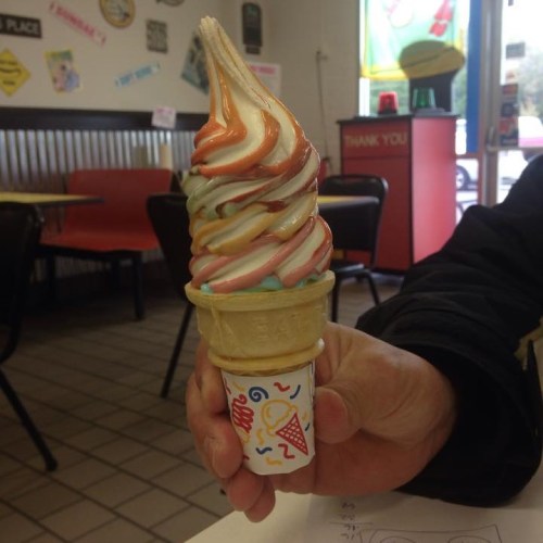 A hand holding a colorful soft serve ice cream cone with swirls of orange, pink, and blue in a paper cup.