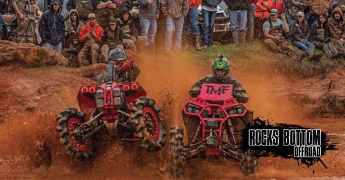 Two ATVs race through muddy terrain, kicking up dirt, with a crowd of spectators watching in the background.