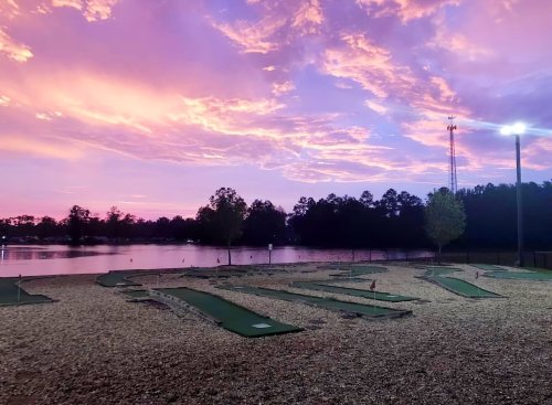 A serene mini-golf course at sunset, with vibrant pink and purple skies reflecting over a calm lake.