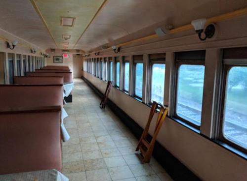 Interior of an empty train car with tables and chairs, featuring large windows and a view of the outside.