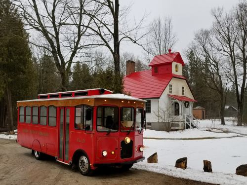 A red trolley parked on a snowy path in front of a white house with a red roof and trees in the background.