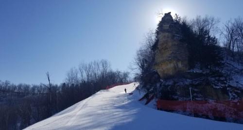 A skier ascends a snowy slope with a rocky cliff and bright sun in the background, surrounded by trees.