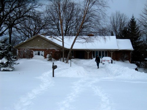 A snowy suburban home with a person walking through deep snow, surrounded by trees and a mailbox in the foreground.