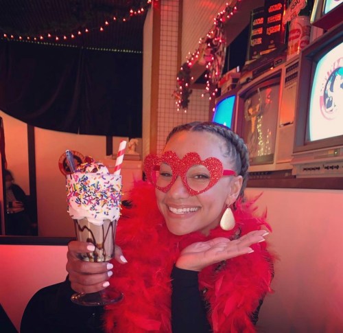 A person wearing heart-shaped glasses and a red feather boa holds a colorful ice cream sundae, smiling playfully.