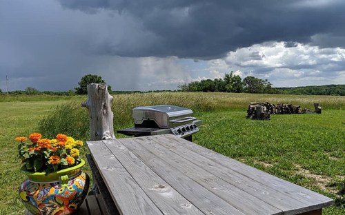 A wooden table with flowers, a grill, and dark storm clouds looming over a grassy field.